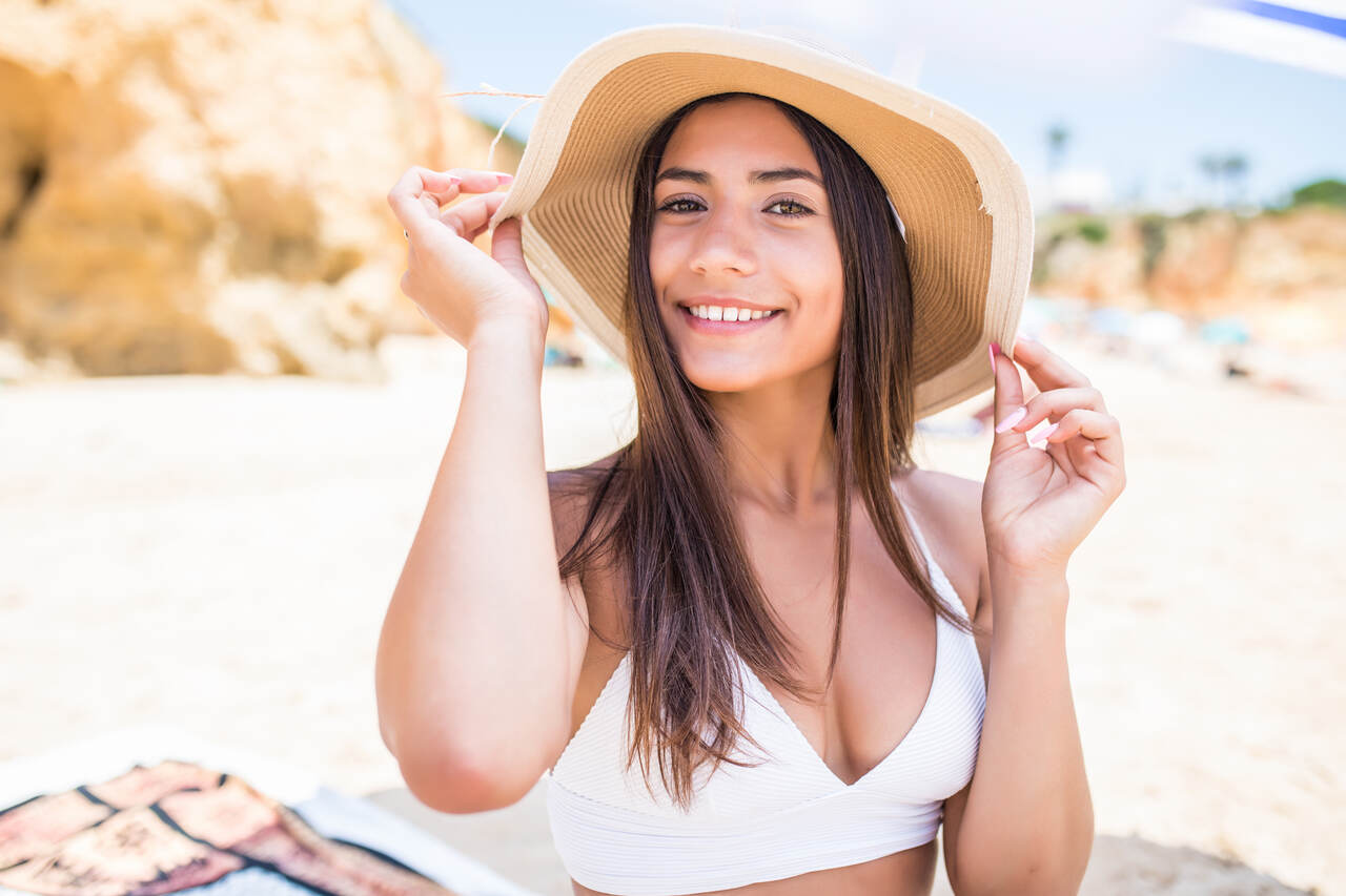 Jovem com belas unhas longas segura seu chapéu sentada na areia da praia 